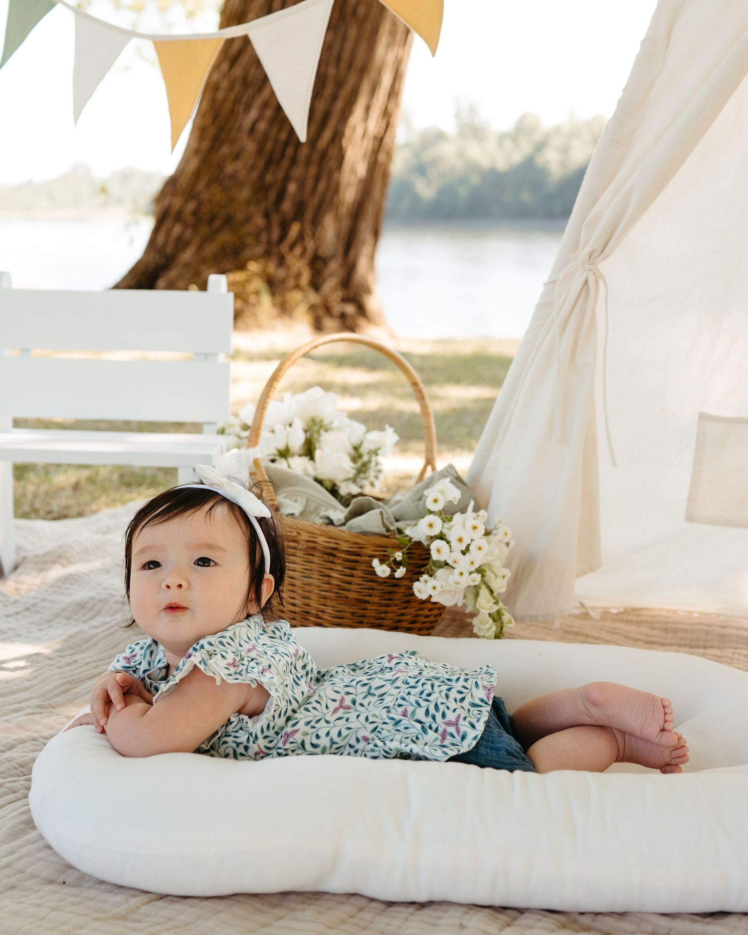 A baby rests on a white cushion next to a picnic basket with white flowers.