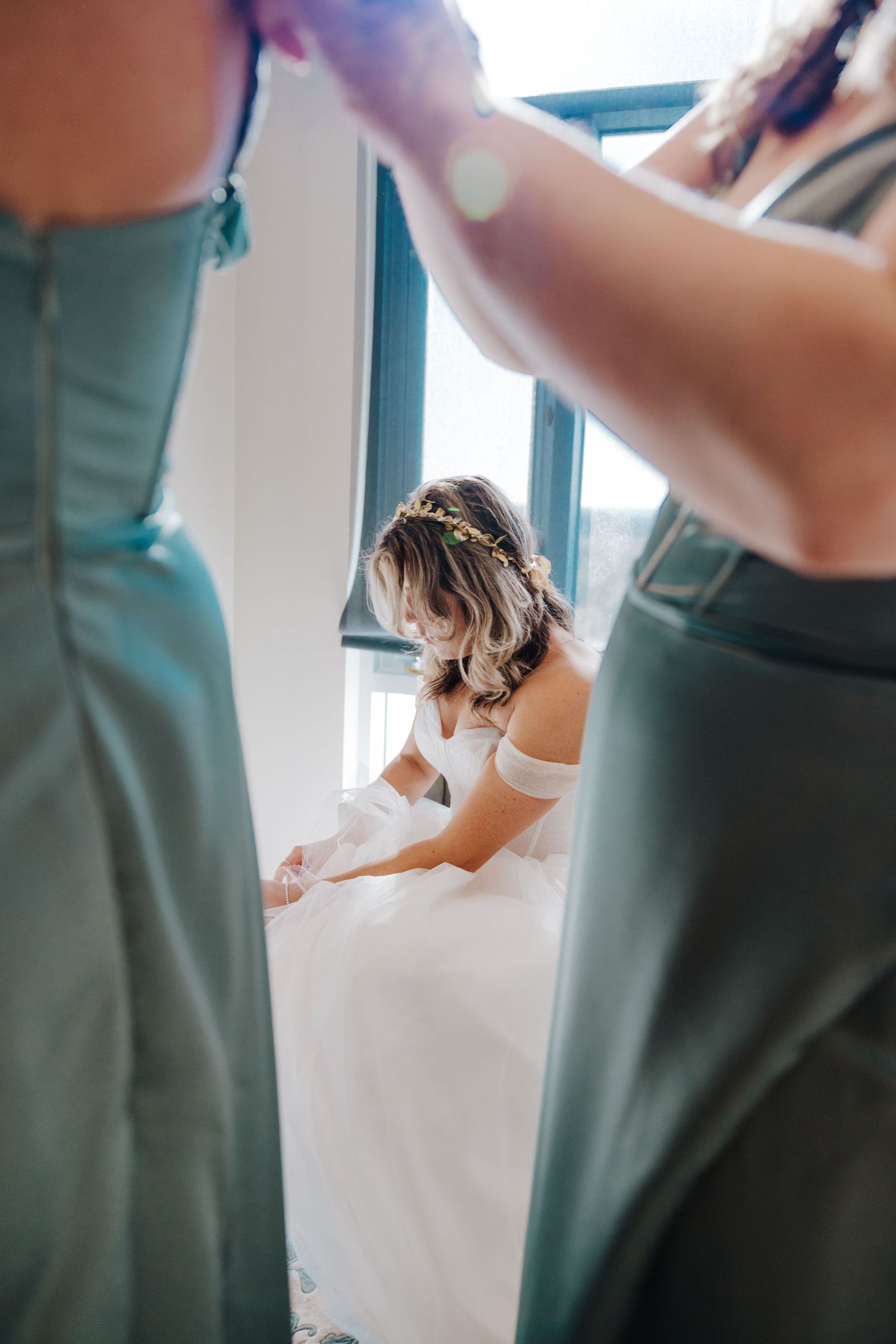 A bride in a white dress sits between two people wearing sage green bridesmaid dresses during wedding preparations.