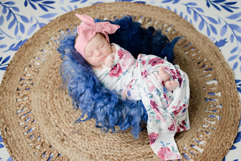 Sleeping baby on navy blue ruffled blanket wearing floral outfit with pink bow resting on woven circular mat.
