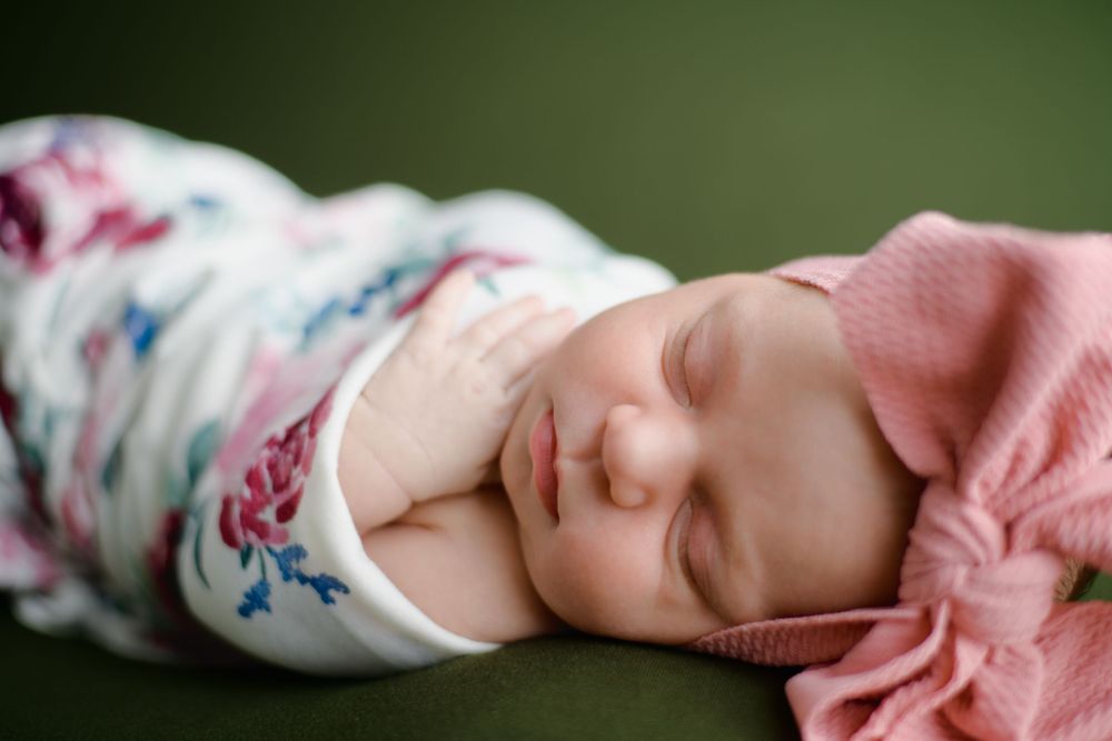 A peacefully sleeping baby wrapped in a floral blanket and pink sweater rests against a green background.