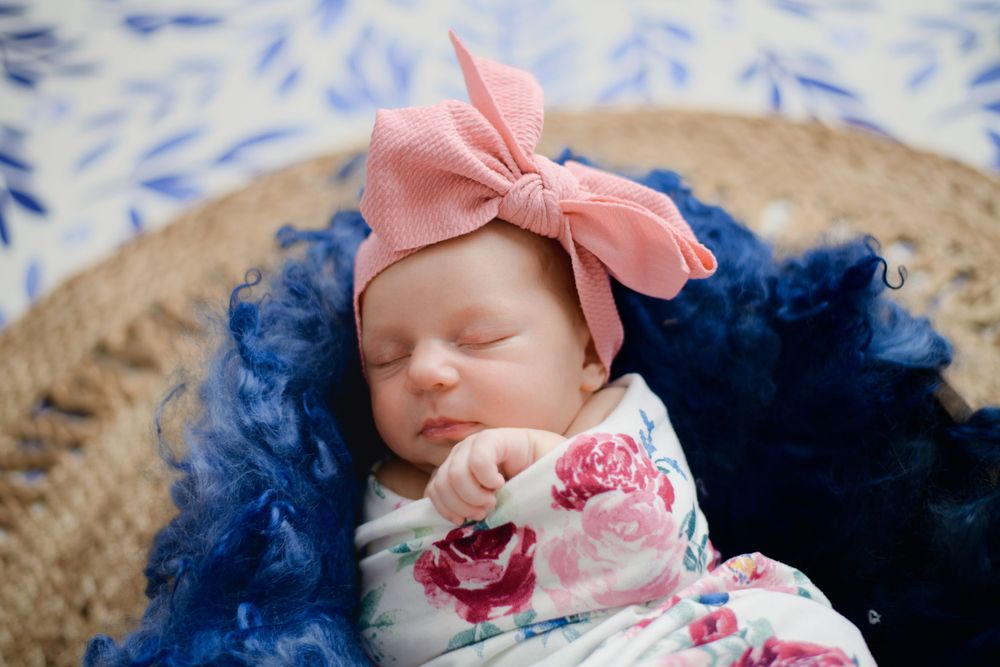 A sleeping baby wearing a pink bow headband and floral outfit rests on navy blue fabric.