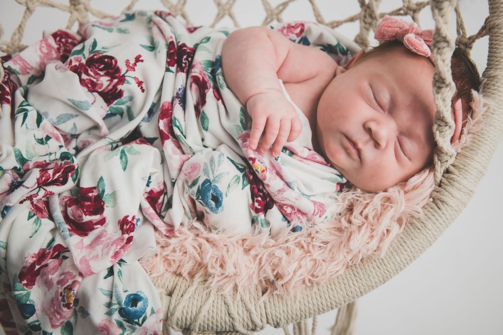 A sleeping baby swaddled in a floral blanket with pink and blue roses lies on a cream fringed pillow.