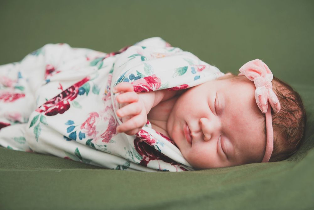 A newborn baby sleeps peacefully while swaddled in a floral print blanket on a green surface.