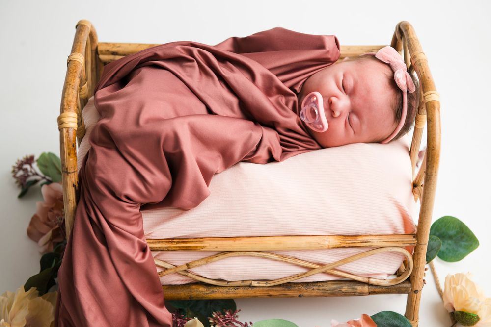 A newborn baby wrapped in a dusty rose swaddle sleeping peacefully in a rustic wooden bed with floral accents.