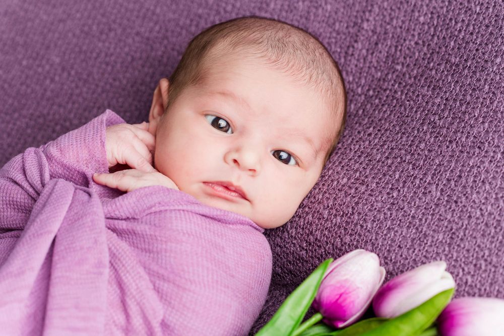 Baby wrapped in purple blanket lies next to pink and white tulips on purple textured background.