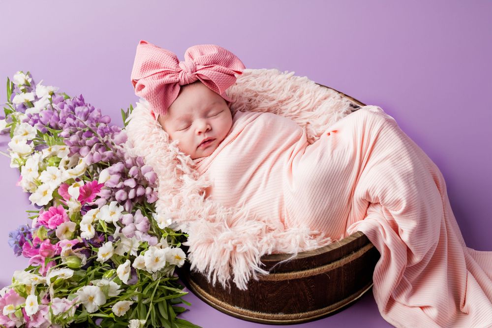 A sleeping newborn in pink wrap lies in a wooden bowl beside colorful spring flowers against a purple backdrop.