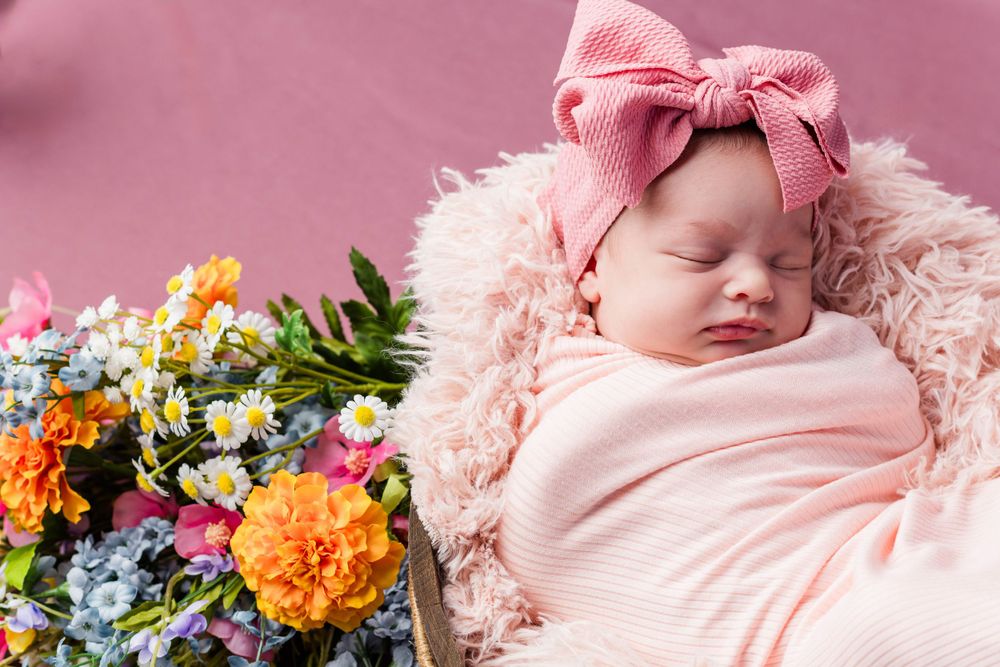 A sleeping baby wrapped in pink swaddle with a pink bow headband lies beside colorful marigolds and wildflowers.