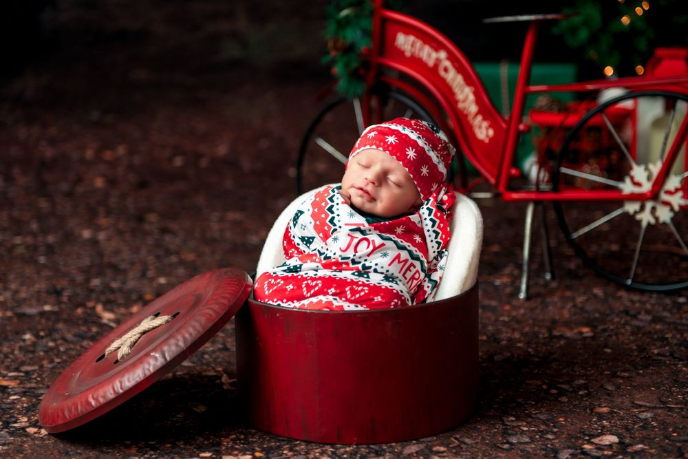 A baby in a cozy red and white Nordic sweater lies in a round red box with Christmas decorations in the background.