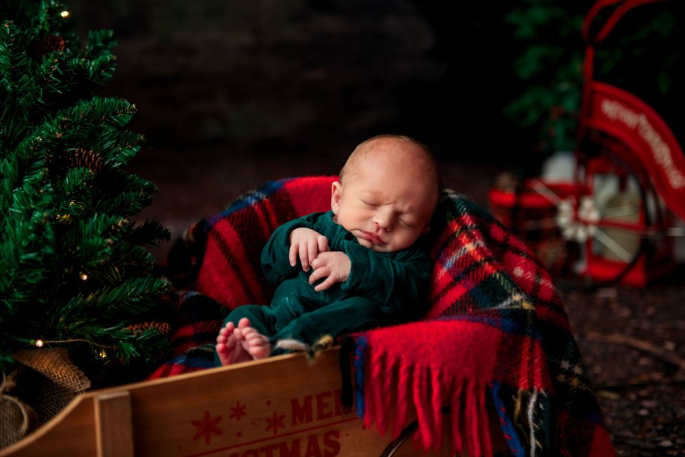 A sleeping baby in festive green attire rests in a wooden box surrounded by Christmas decorations and plaid blanket.