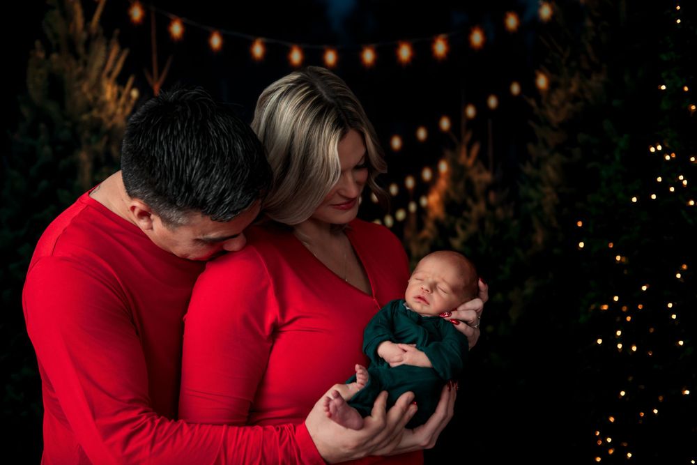 A couple in red sweaters holds their newborn baby under glowing string lights during a holiday photo session.