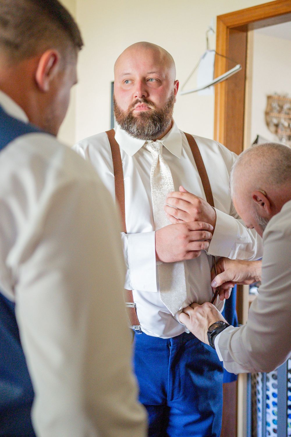 Bald bearded groom in white shirt with suspenders gets help from groomsmen adjusting his tie before wedding.