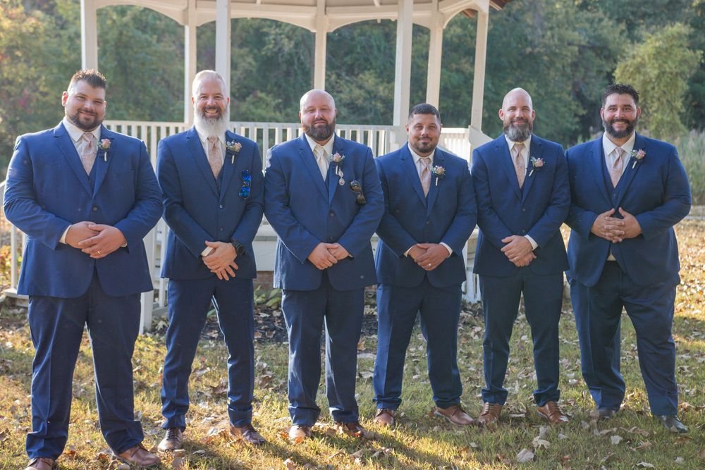 Six groomsmen in navy blue suits with tan ties pose outdoors near a white gazebo on a sunny wedding day.