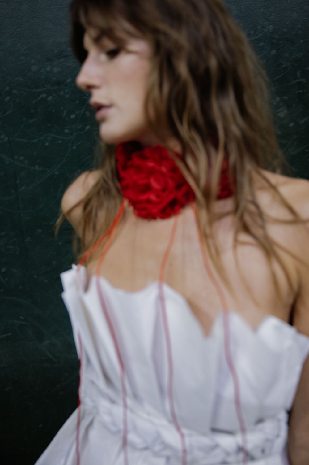 A figure in a white dress with tousled brown hair and a dramatic red rose collar poses against a dark background.