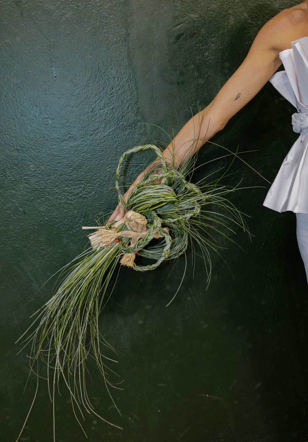 Hands holding and manipulating a bundle of dried grass stalks against a dark green wall background.