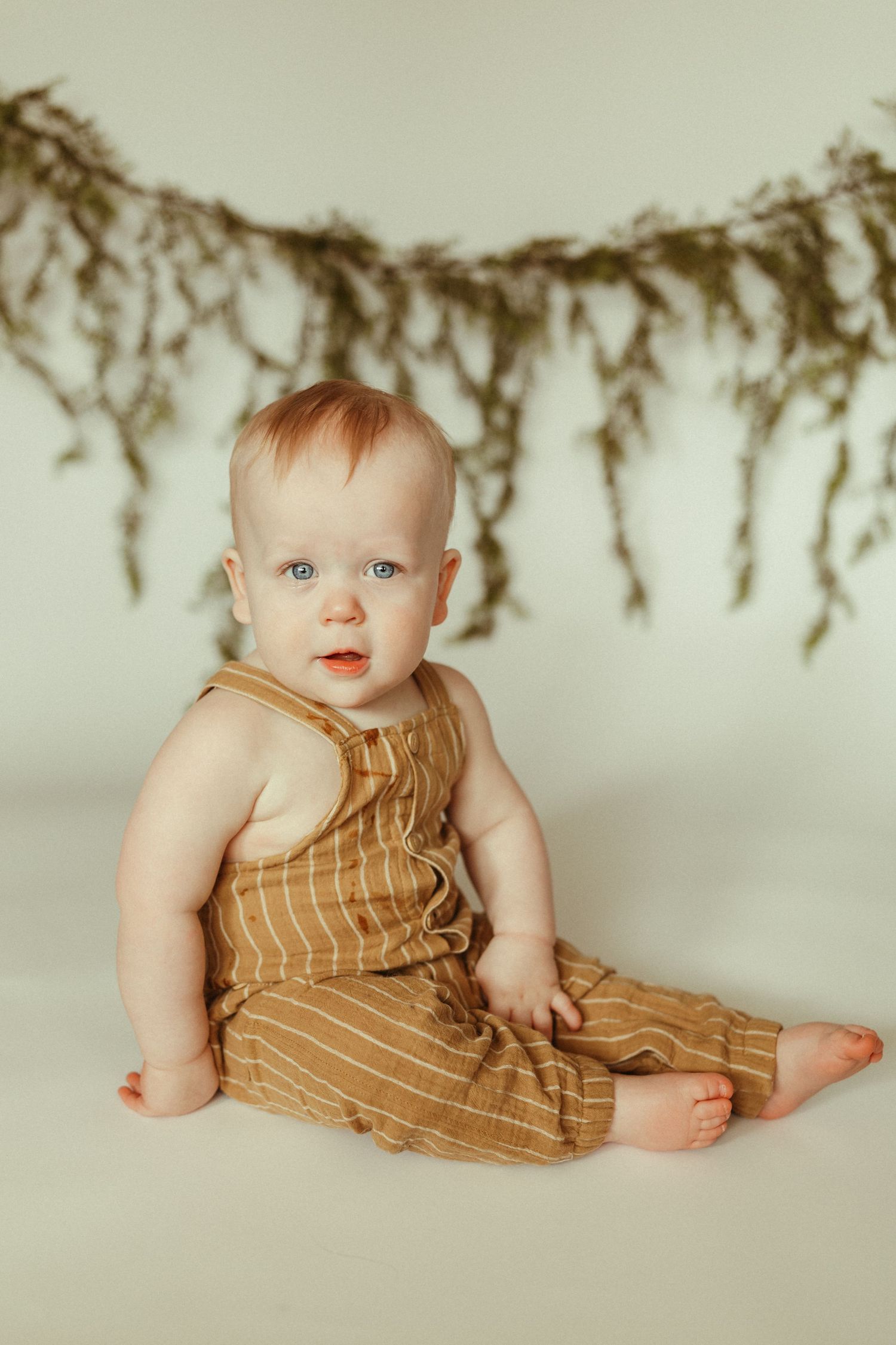 Baby wearing brown striped romper lays on white floor with dried flowers hanging above in studio setting.