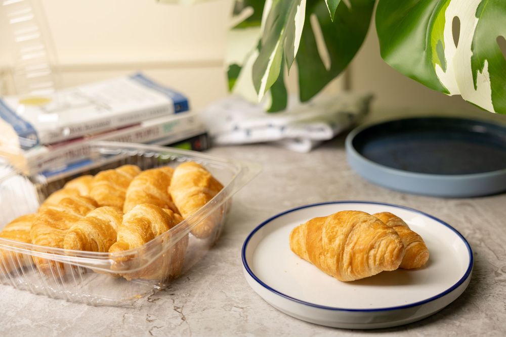 Fresh baked croissants on a white plate with blue trim, with more pastries in a plastic container in the background.