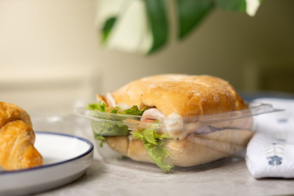 A freshly made sandwich with deli meat, lettuce and cheese on a ciabatta roll sits on a clear glass plate near a croissant.