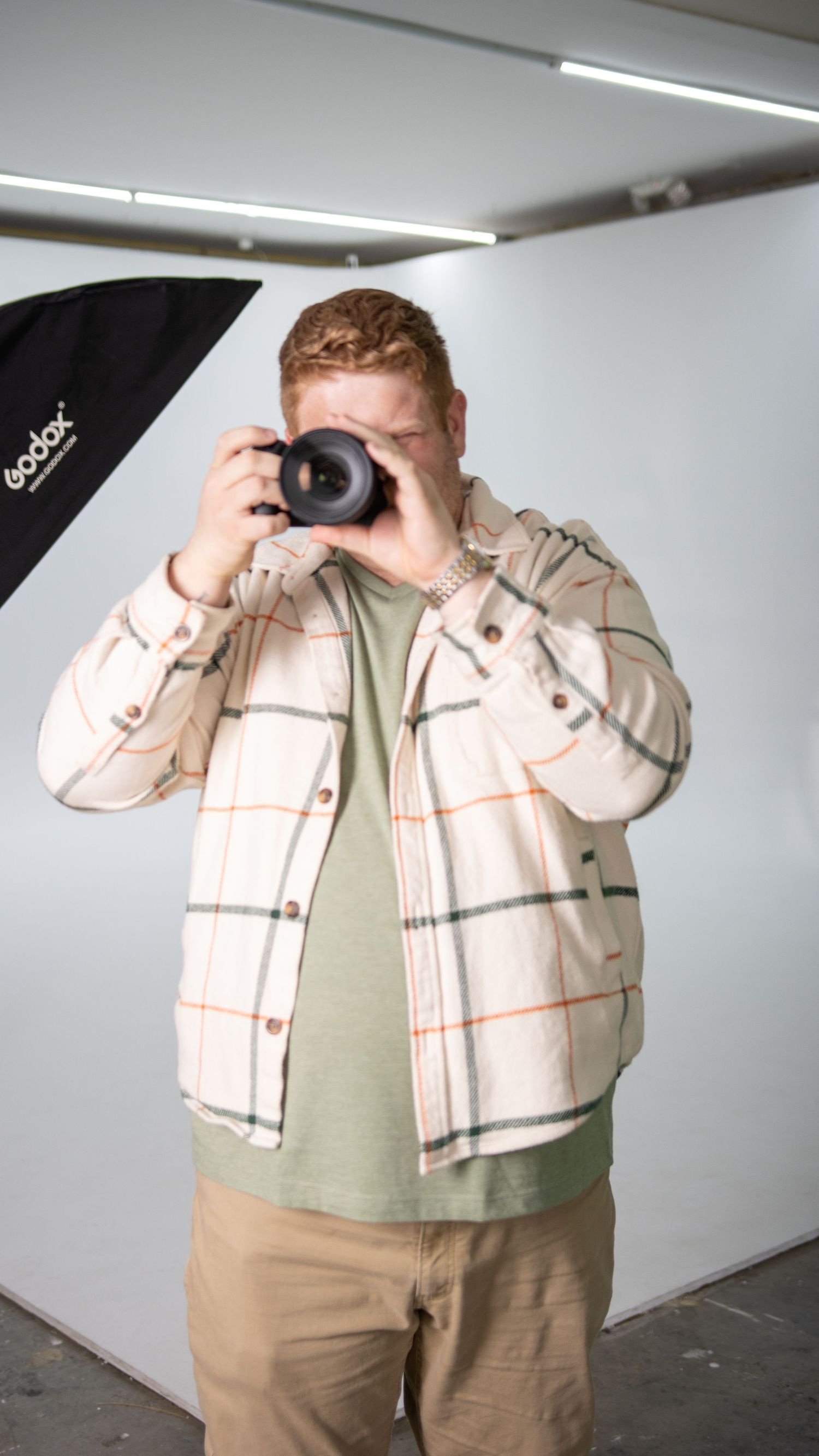 Person in beige clothing positioning a camera in front of their face in a studio setting with white walls.