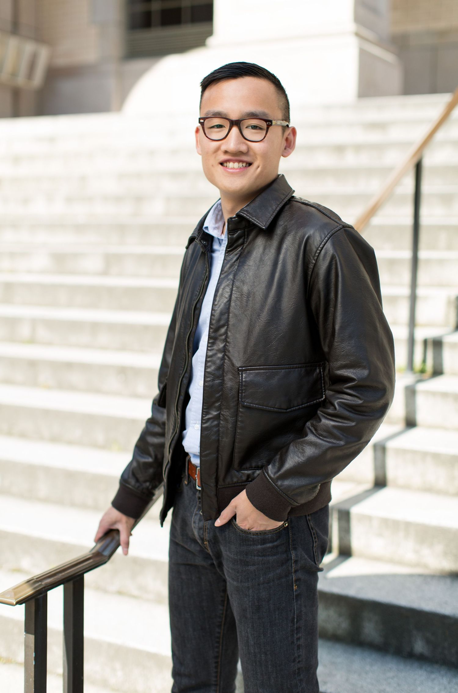 A person in a leather jacket stands smiling on concrete steps.