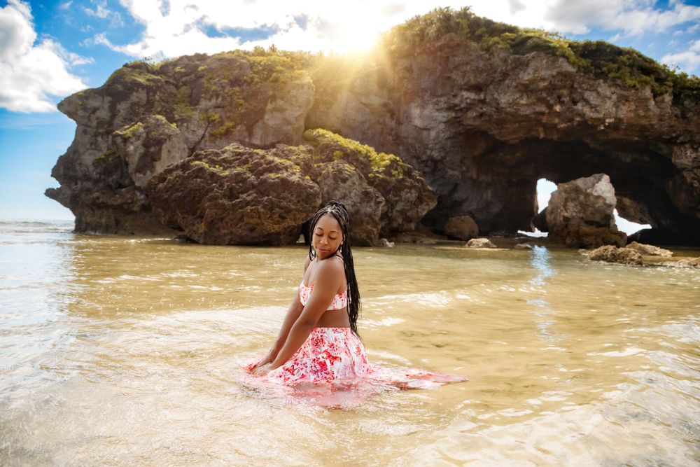 Someone in a pink dress poses in shallow ocean water near a dramatic rock formation.