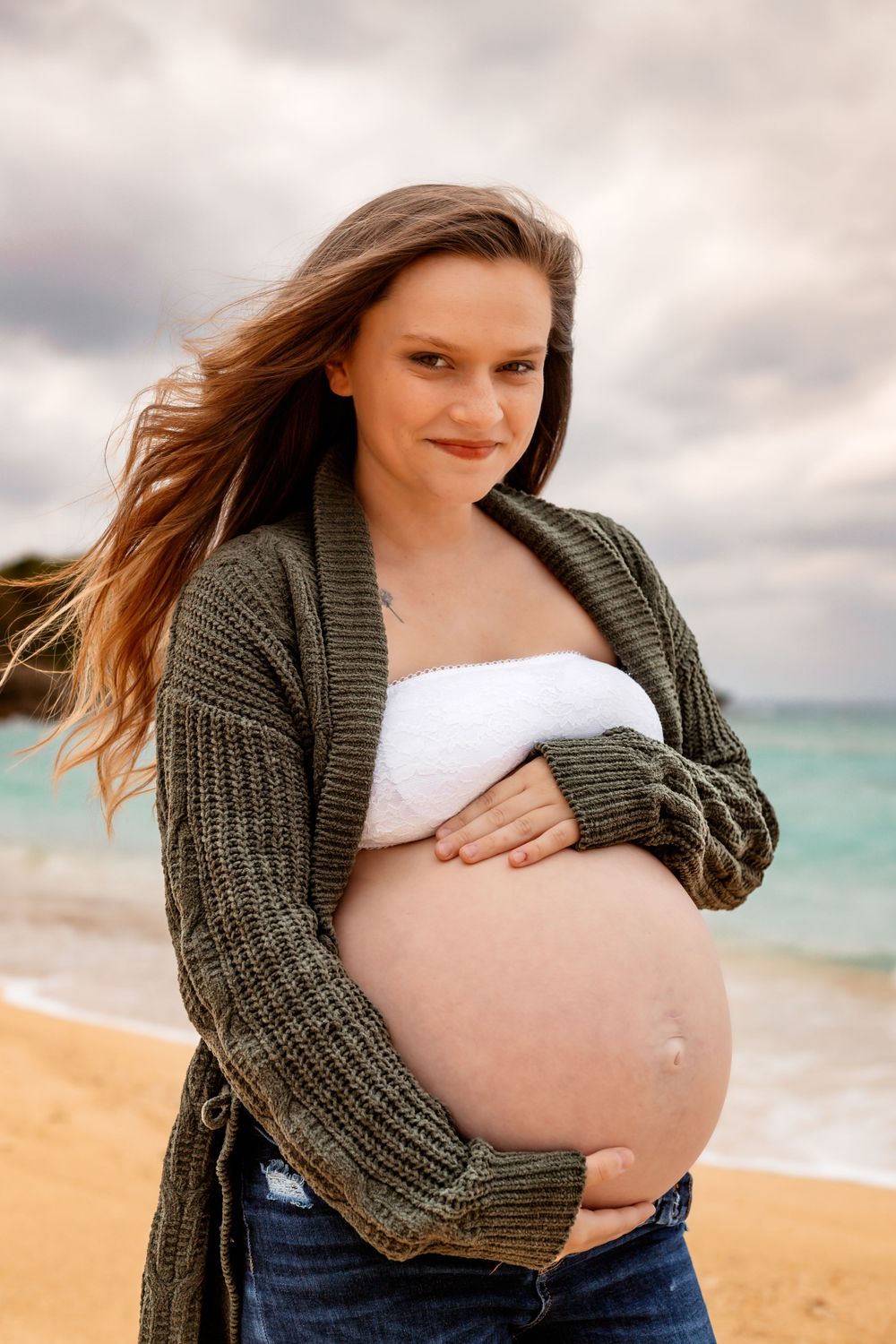 Expectant mother in knit cardigan stands on beach during sunset.