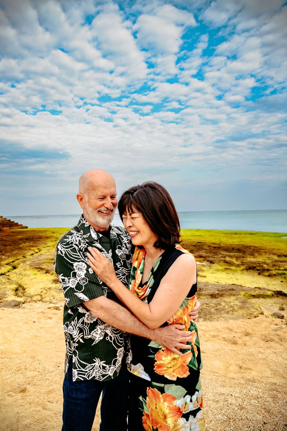 A loving couple laughing together on a coastal cliff wearing matching floral Hawaiian shirts.