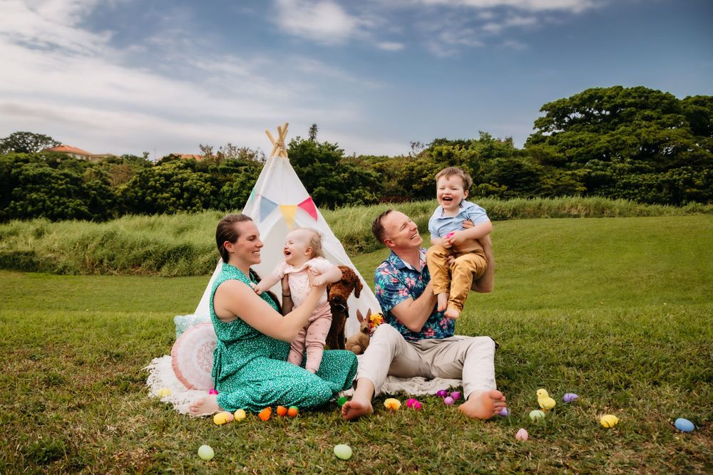Family picnic scene with teepee tent and scattered Easter eggs on green grass under blue sky.