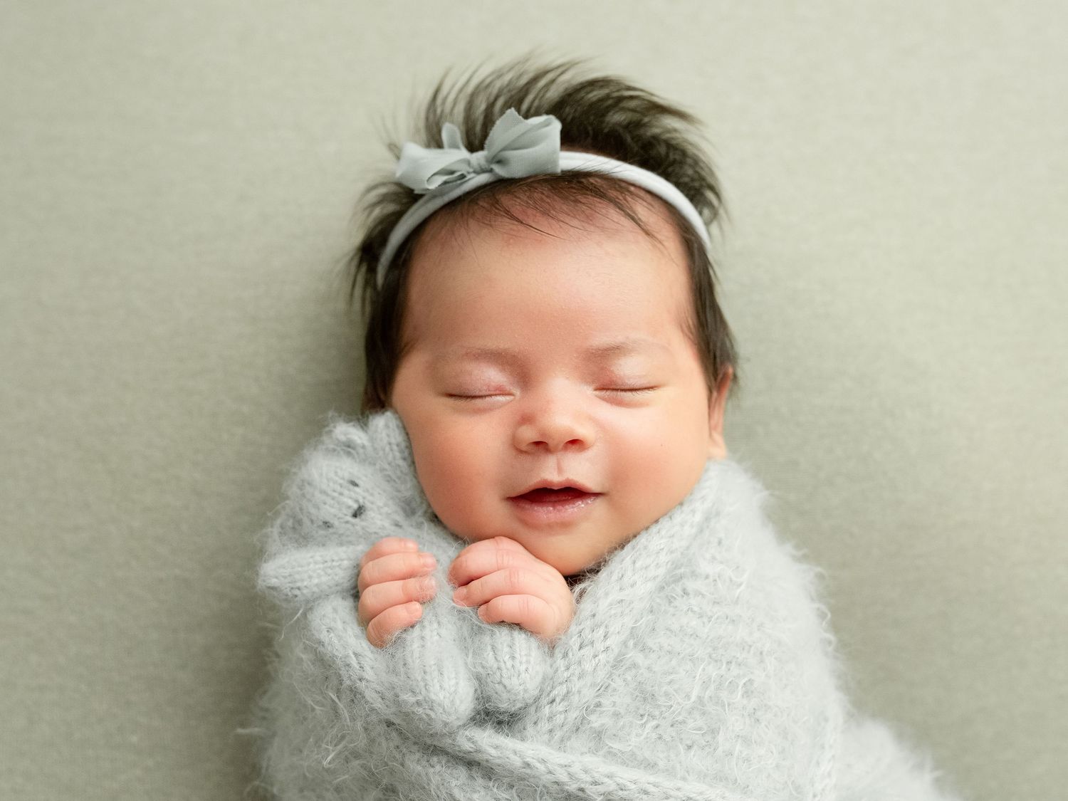 A peaceful sleeping newborn baby wearing a delicate white bow headband lays on a neutral background.
