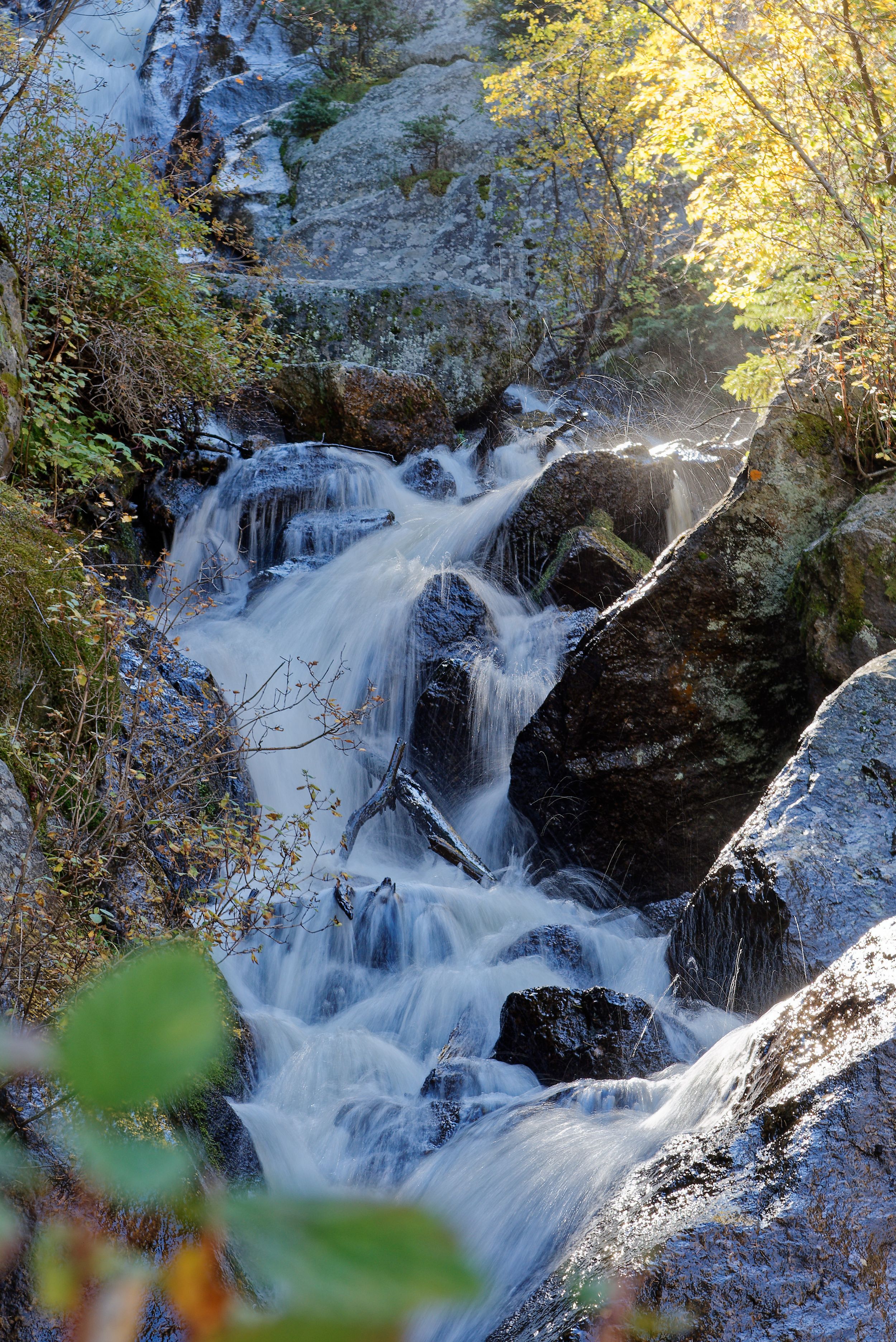 Cascading waterfall flowing over rocky terrain with autumn foliage and sunlit trees in the background.