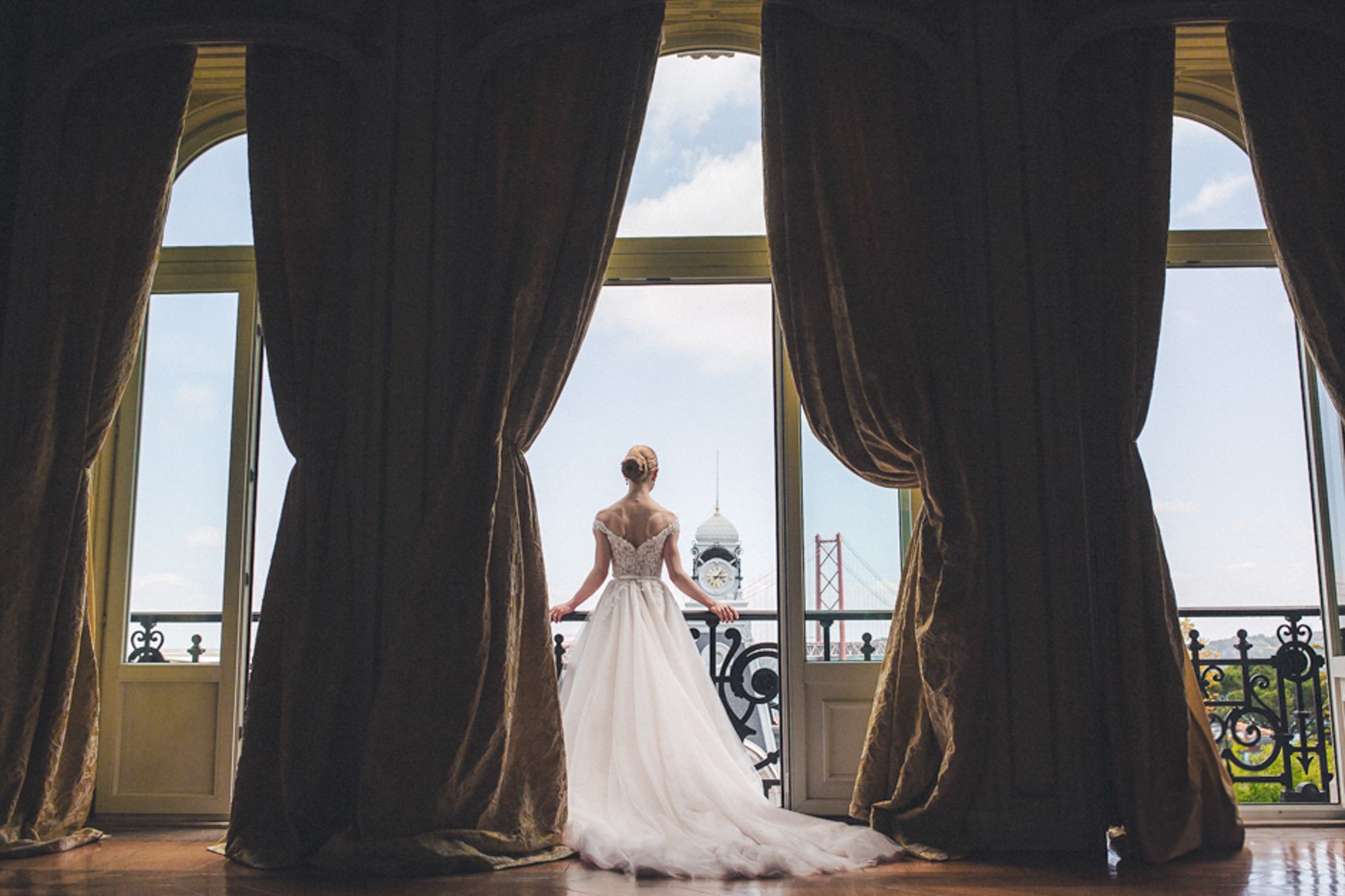 A bride in a blush wedding dress stands at a window at Lisboa Pestana palace. Captured by a destination wedding photographer in Portugal