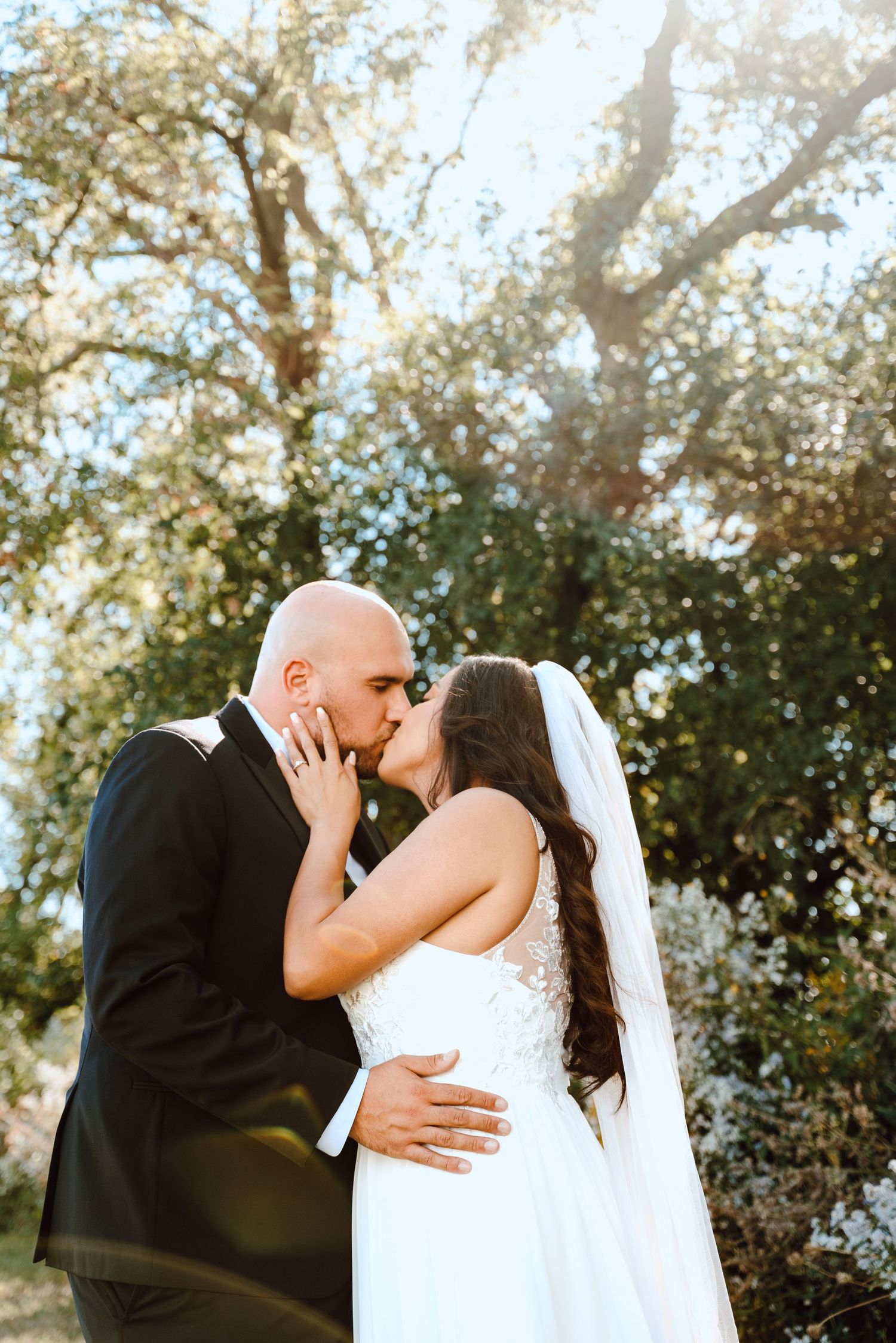Beautiful outdoor wedding scene shows a tender kiss between newlyweds surrounded by sunlit trees during golden hour.