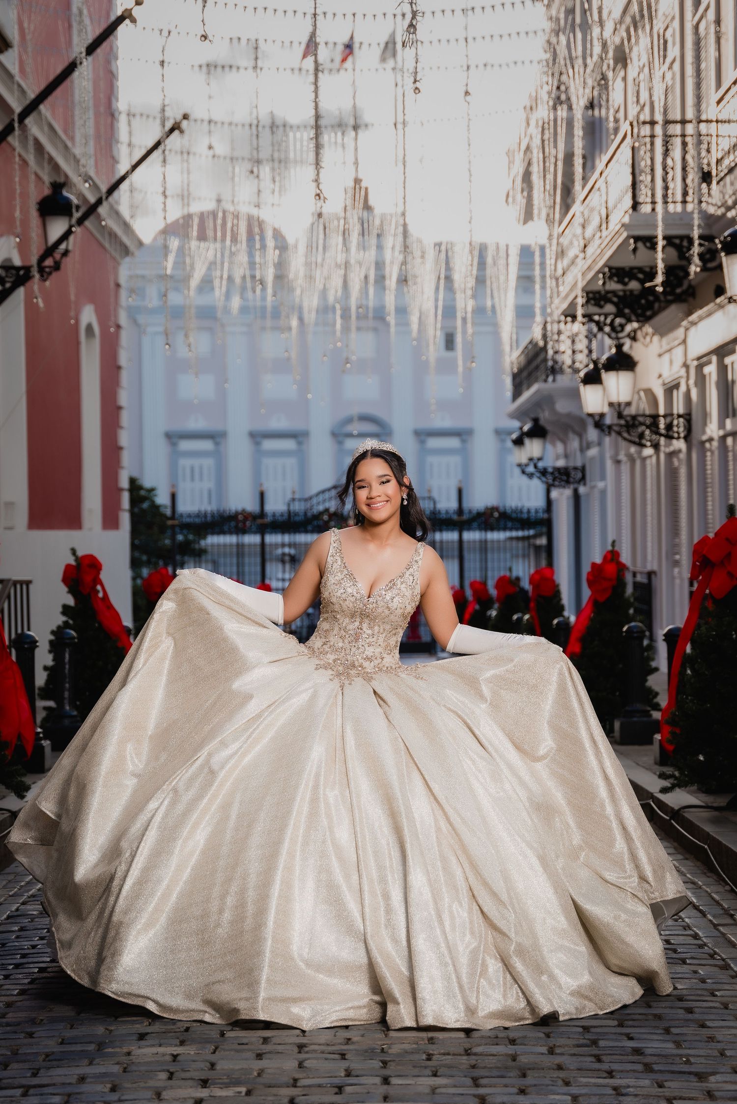 A wedding gown with a sparkly bodice and full ballgown skirt is displayed on a cobblestone street with red flowers and lights.