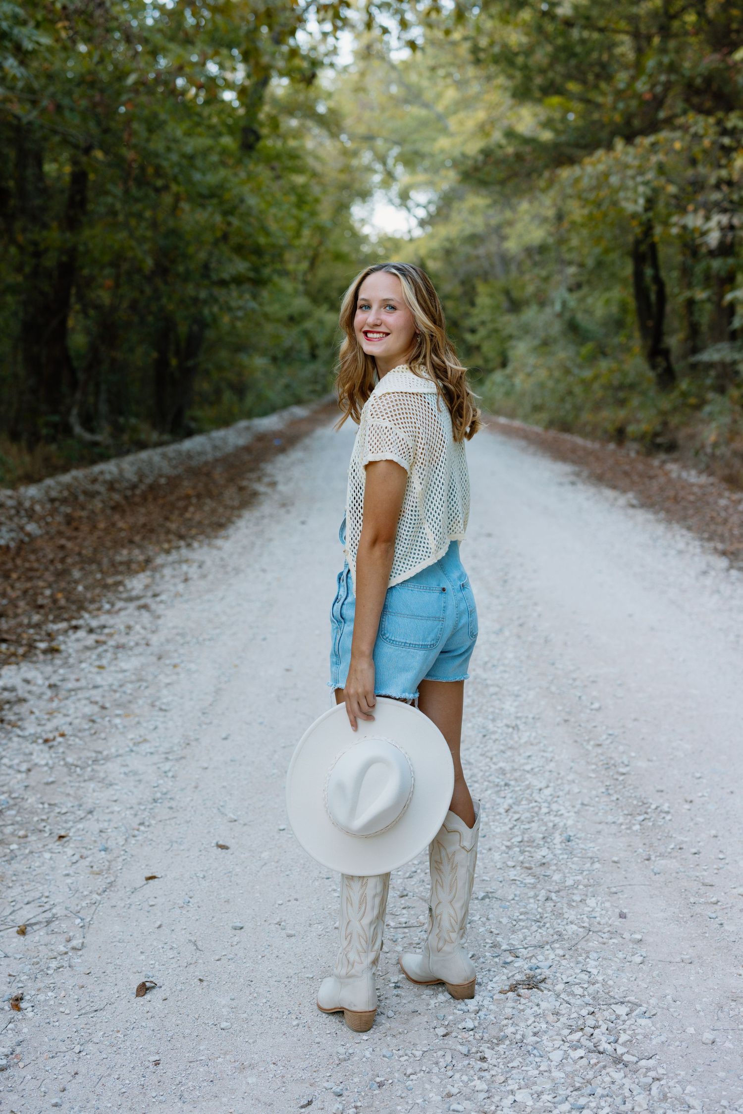 Woman in cowboy boots and shorts poses on dirt road holding white hat.