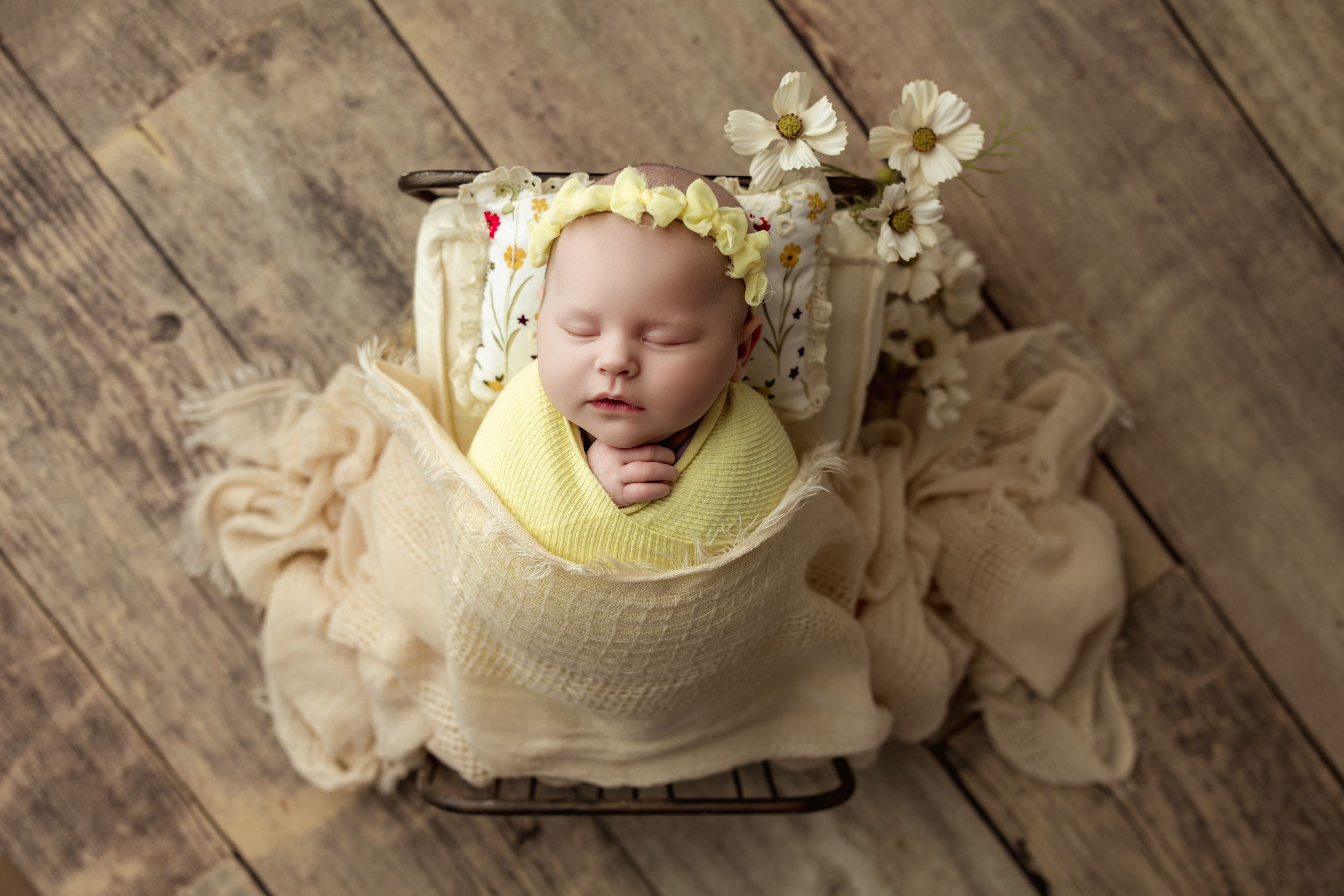 Newborn baby wrapped in yellow swaddle with floral bonnet sleeping in a rustic wooden bed.