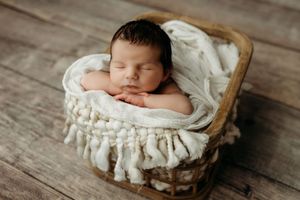 Peaceful newborn baby sleeping in a rustic wicker basket wrapped in a soft white knit blanket.