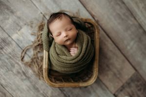 Newborn baby swaddled in olive green wrap sleeping peacefully in a rustic wooden bowl on a gray wood floor.