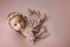 Newborn baby girl in pink outfit and floral headband lying on pink backdrop in professional portrait.