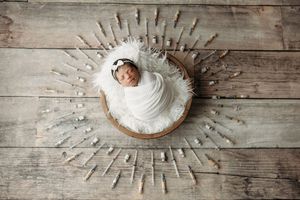 Newborn baby swaddled in white wrap sleeping in wooden bowl surrounded by decorative sticks on rustic wood floor.