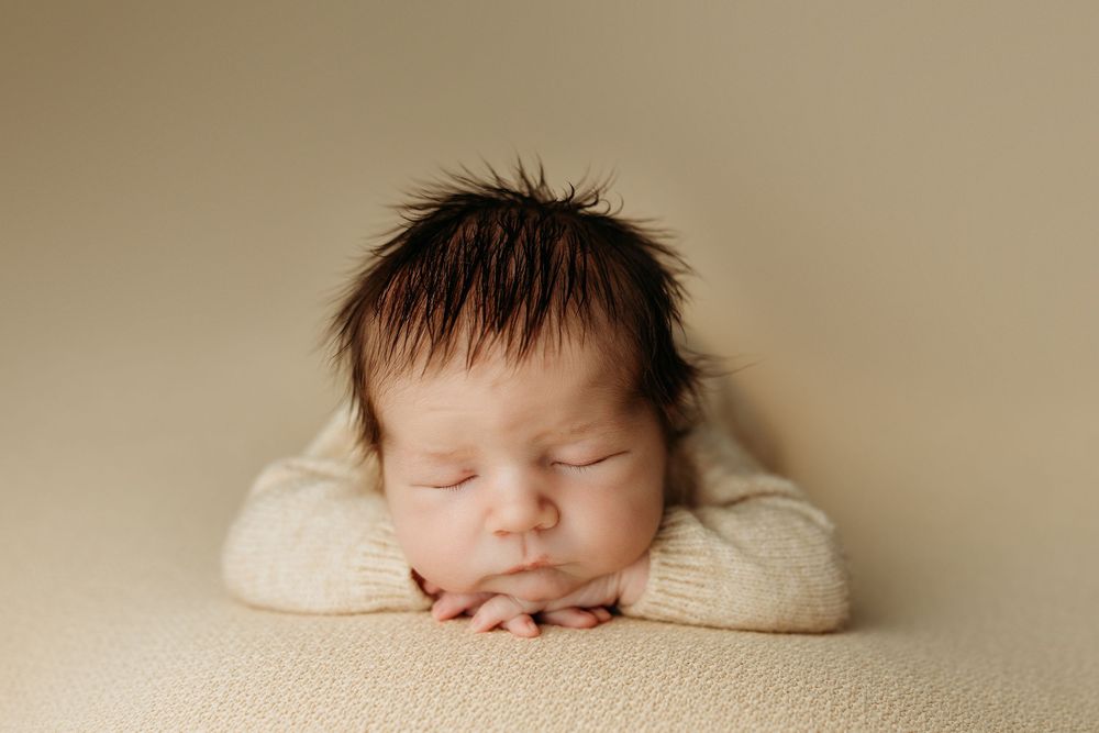 Sleeping newborn baby with dark spiky hair resting chin on hands, wearing cream knit outfit on beige background.