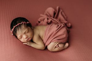 Sleeping newborn baby girl wrapped in dusty rose fabric with floral headband on matching pink textured backdrop.