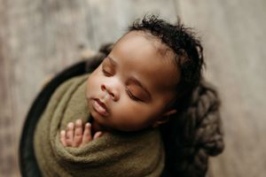 Peaceful newborn baby sleeping wrapped in olive green swaddle, nestled in wooden bowl, professional newborn photography.