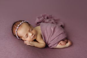 Sleeping newborn baby girl wrapped in a purple swaddle with a floral headband on a mauve background.