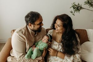 A couple sits on a brown sofa, smiling at their newborn baby dressed in green, in a bright, minimalist room.