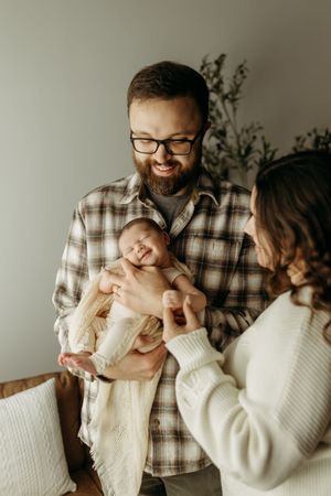 Smiling bearded dad holding newborn baby while mom in white sweater looks on lovingly indoors.