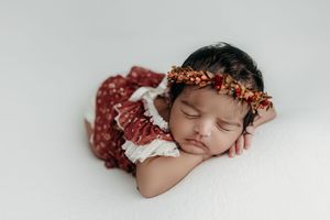 Sleeping newborn baby girl in red floral romper and matching flower crown headband on white backdrop.