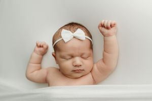 Newborn baby girl sleeping peacefully with arms raised, wearing a white bow headband on a soft white background.
