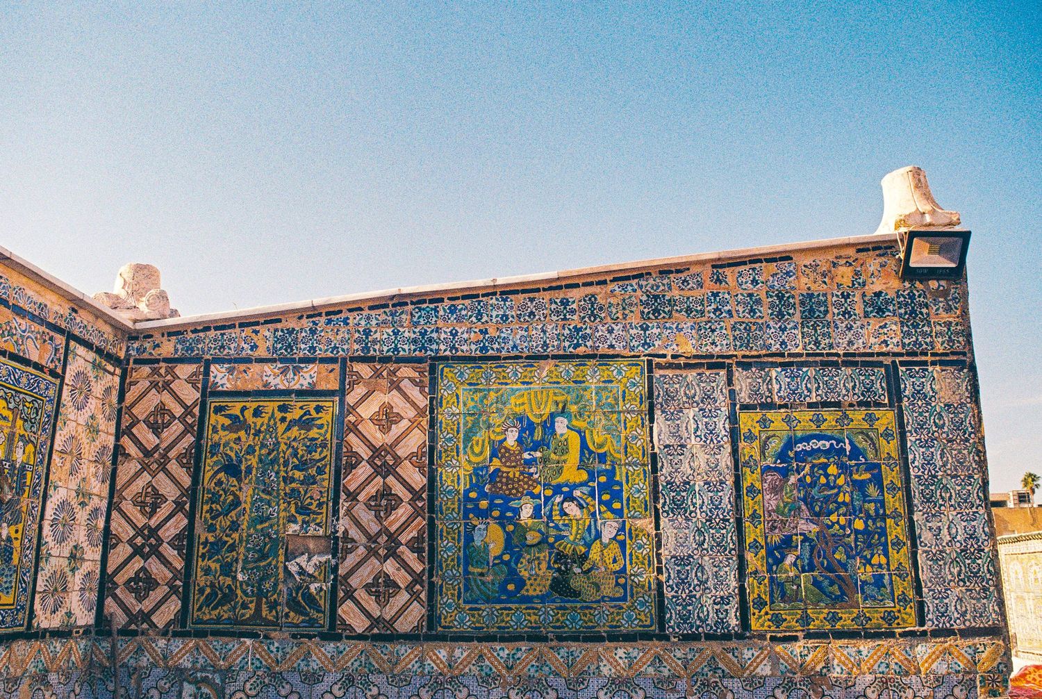 Ornate mosaic tiles with geometric patterns and blue and gold details decorate an ancient wall against a bright blue sky.