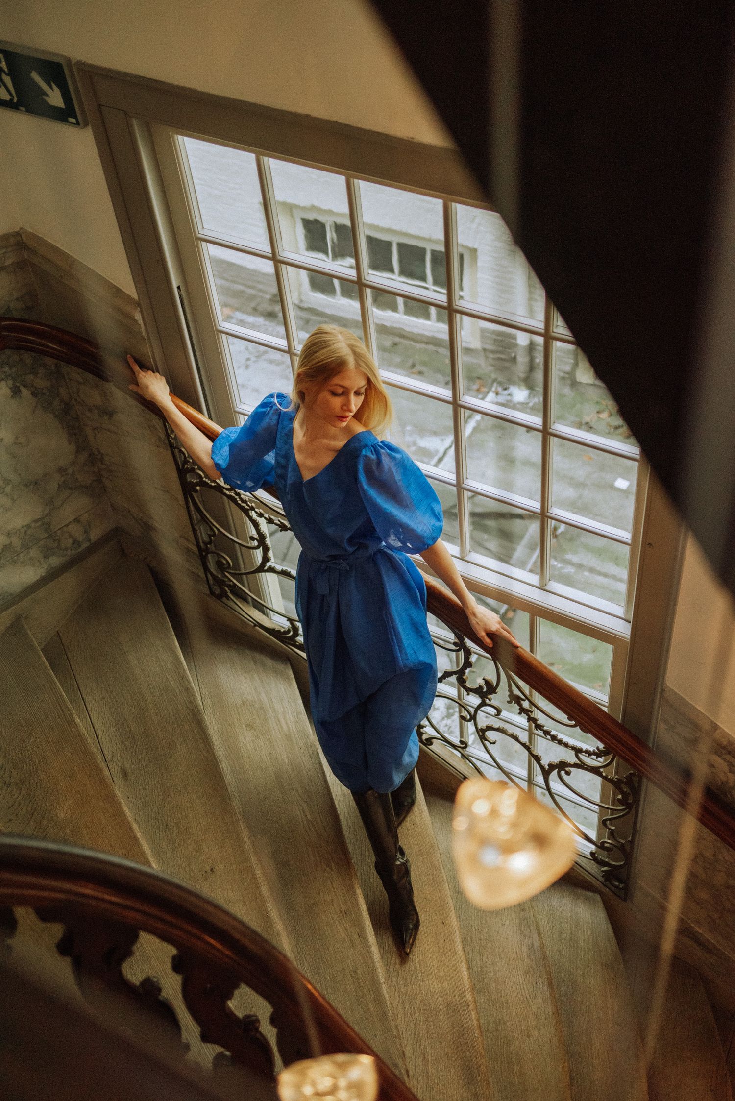 A woman in a blue dress poses on an elegant staircase with dramatic window lighting.