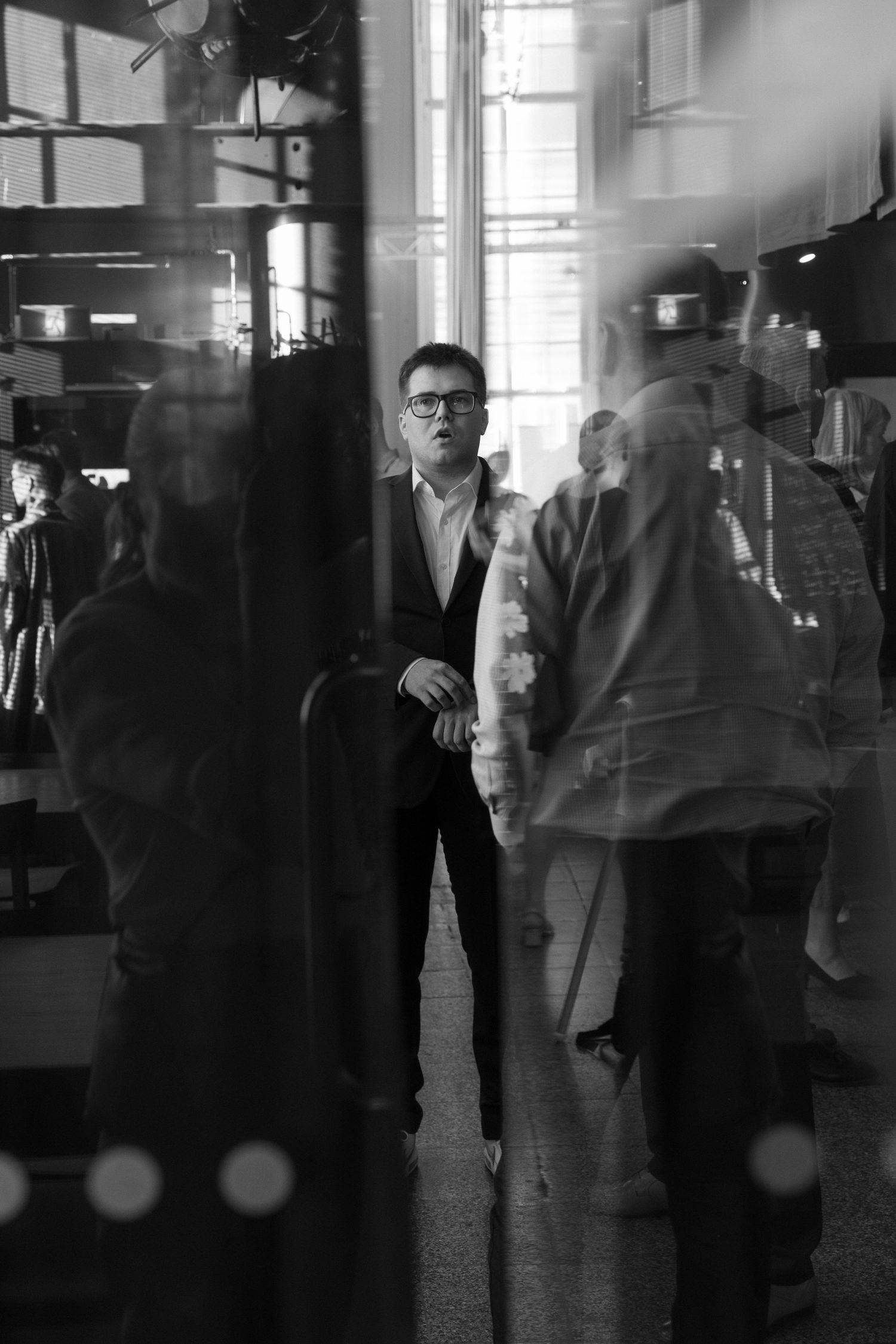 Mustavalkoinen valokuva, jossa hahmot heijastuvat lasiin ja luovat kerroksellisen, tunnelmallisen kuvan juhlista.