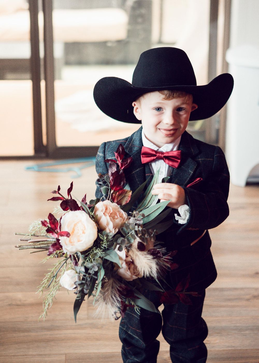 Little boy in black cowboy hat and bowtie holds a rustic burgundy and cream floral bouquet against wooden backdrop.